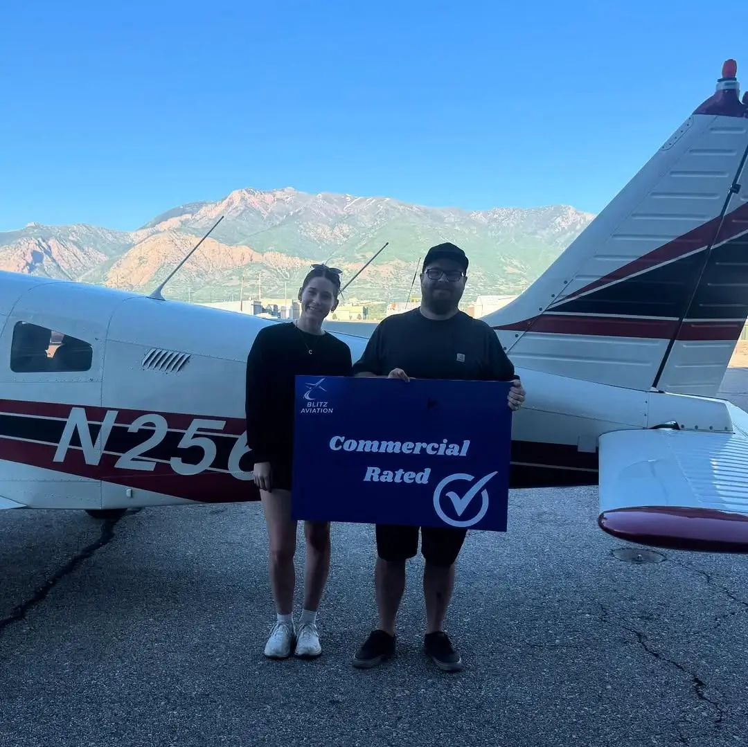 Student and CFI standing in front of and airplane while holding a poster with "Commercial Rated" on it