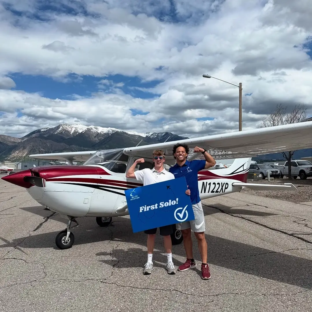 Student and CFI smiling and flexing their arms in front of an airplane while holding a poster with "First Solo" written on it